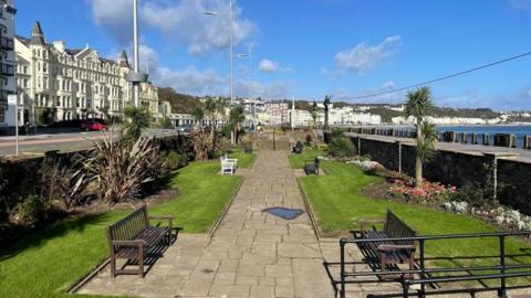 Well maintained sunken gardens with a footpath and benches along Douglas Promenade. Tall pale coloured buildings can been seen along the road side on the left, with the sweeping blue curve of Douglas Bay on the left. A bright blue sky above features some white clouds.