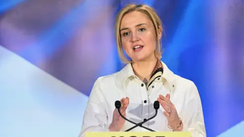 Mairi Mcallan talking at a yellow podium with two mics