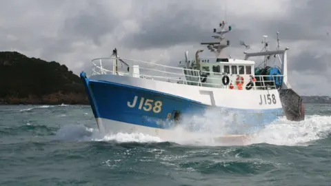 States of Jersey Police A white and blue fishing boat with J158 on the left side, in the ocean, next to another vessel out at sea with grey skies and land seen in the background.