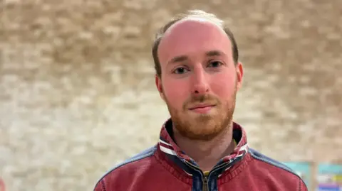 Rory Clark looking into camera with a brick wall behind him inside a community centre. He has short hair and a closely shaved beard and is wearing a red zip-up jumper with a blue trim near the zip.