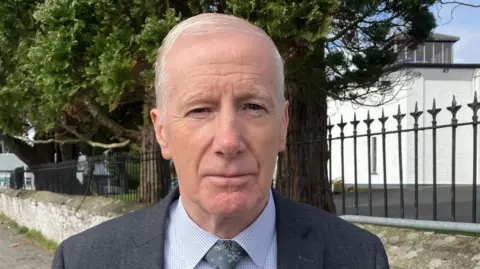 Gregory Campbell is standing outside near a number of trees and a black fence. He is wearing a grey suit, green tie and blue shirt. 