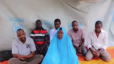 AFP A still taken from a video showing a kidnapped aid workers, and five men believed to be her colleagues. They are all sitting on mats in front of tarpaulin.