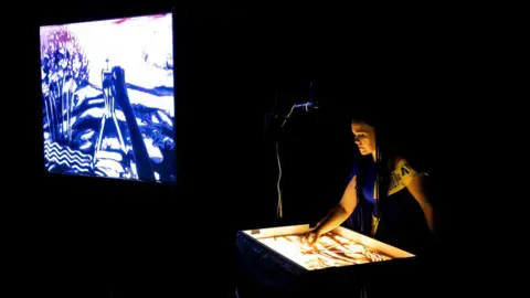 Olena Kalinina  A woman projects art onto a large screen from her own illuminated working station, which has a light covering of sand. She is at the Trinity Centre in Bristol and is carrying out her performance in the dark