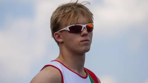 A young man with blond hair wearing white sunglasses and a white Jersey. Behind him is blue sky with white clouds. 