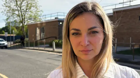 Joanna Wade with long blonde hair, looking at the camera and wearing a white shirt. There is a single-storey brick court building behind her, with a railing around the roof. There are concrete bollards along the pavement outside, and a narrow road behind Ms Wade, with double yellow lines.