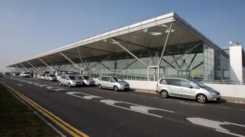 The drop off area outside stansted airport, with several taxis parked up. It's a clear, sunny day.