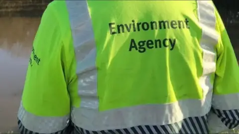 The back of an Environment Agency worker wearing a fluorescent yellow high-visibility jacket with reflective strips and the words “Environment Agency” printed on it.