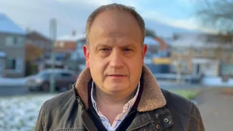 Councillor Dan Morris on a residential road, wearing a brown coat with snow on the ground behind him. He has short, light-brown hair and is clean shaven. A blurred image of a car next to a green verge is behind him with buildings, blurred, in the distance beyond