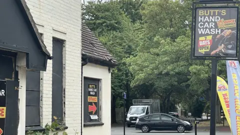 A picture of an abandoned white pub to the left with black boarded up windows. To the left a black car and a white van can be seen in the car park. A Butt's Hand Car Wash sign can also be seen