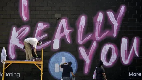 Getty Images Three men spray-paint a Facebook dating advert which reads "I Really (Facebook like icon) you"