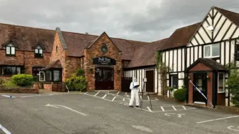 The Pear Tree Inn. A red-brick building with an officer in forensic PPE and police tape sealing off a black door into the building.