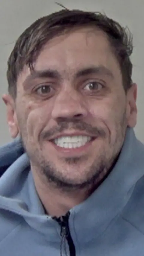 A man in a blue tracksuit with white teeth in a police mugshot.