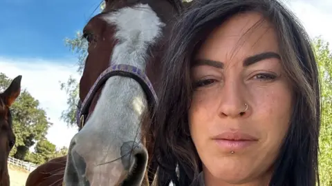 Thea Creasy takes a selfie while standing next to a horse. Ms Creasy has long dark hair and has a nose ring. The horse is a bay colour with a white section stretches from its forehead to its nose.