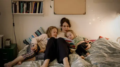 Getty Images Mother reading picture book while sitting with children in bedroom - stock image