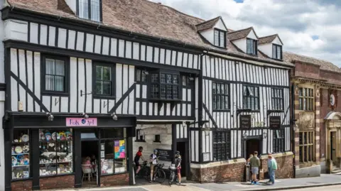 Ricky Barnett A black and white Tudor-style building, part of which is over a Pots of Art shop. There are three cyclists outside and some other members of the public.