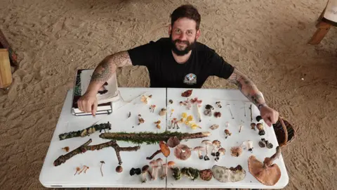 Bere Marsh Farm A bird's eye view photo of Andy Knott. He is looking up and smiling for the camera. There is a variety of mushrooms, tree branches and books on a white table in front of him and a basket on the floor beside him. He is wearing a black shirt and has tattoos on his arms. The ground is covered with sand.