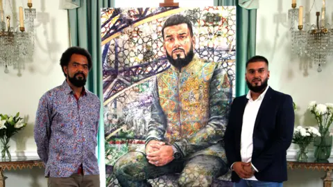 Jack, left, and Rizwan stand beside a vibrant, mosaic-inspired portrait of Rizwan in an elegant room with chandeliers and floral arrangements.