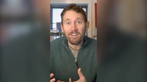 A man with short brown hair, a moustache and beard talks to the camera. He is wearing a green jumper with an unzipped collar.