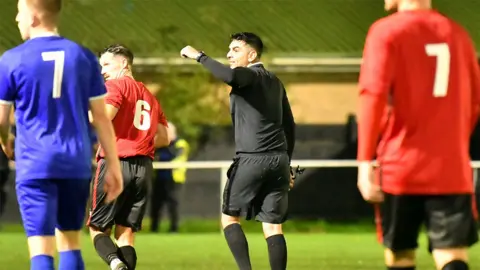 Geoff Quinn Rohan, an Asian man, wearing a black top, shorts and socks as a referee. He is looking to his left with an arm raised, and around him are players wearing blue and two players wearing red. They are on a green pitch.