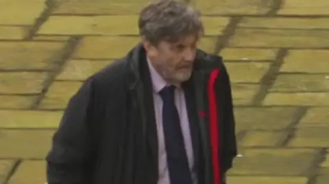 Alan Harris walks towards Winchester Crown Court. He is wearing a black coat which is unzipped and a shirt and tie. He has short greying dark hair and a greying beard.
