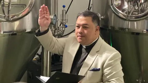 The Dean of Gloucester Cathedral, perhaps in his early 50s, wearing a cream suit and a dog collar, raises his hand as he stands in front of vats of beer, blessing it