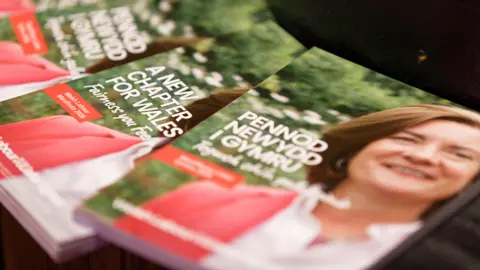 A set of manifestos laid out of a surface with Eluned Morgan's face on the booklets. The manifestos says "Pennod Newydd i Gymru" and "A New Chapter for Wales".