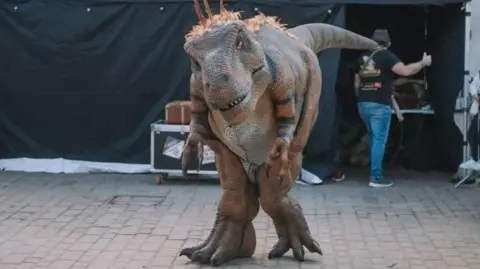 South Kesteven District Council Dinosaur puppet stood on paving in front of a black curtain, a box of props and a man with dark hat, dark top, blue jeans and trainers. 