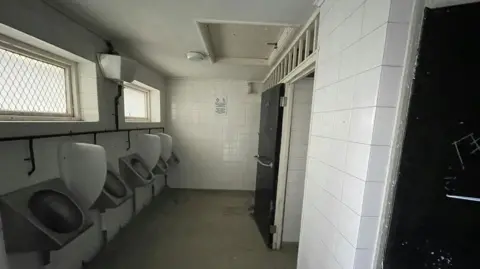 Dyer & Hobbis Five metal urinals on a wall on the left of the image. There are two rectangle windows above them. The walls are white tiles and the floor is light brown tiles. there are two black doors on the right wall