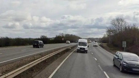 Part of the A14 close to Titchmarsh in Northamptonshire. The road has two lanes on each carriageway and a number of cars and vans can be seen travelling in both directions. There are trees and bushes at the sides of the road. The picture is taken in daylight and the sky is cloudy