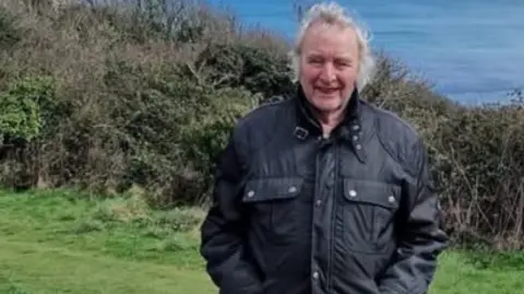 A man wearing a coat who has long grey hair. He is standing on a grass bank in front of the sea, which can be seen behind him.