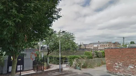 There is a paved area in the foreground with a brick wall on the right hand side and a tree on the left hand side. There is street lighting and a telegraph pole with a large building in the distance.
