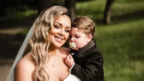 Family A woman with long blonde hair in a wedding dress with a long white veil is holding her infant/toddler son in her arms. The  boy is wearing a black suit jacket and has a white dummy in his mouth.