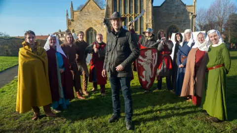 Visit Worcestershire A group of people stand on grass in front of an old stone church, dressed in medieval-style clothing and armour. In the centre, a man in modern outdoor clothes and a hat smiles at the camera, while others around him wear long robes, cloaks, helmets, and carry swords and a red shield with a white lion emblem.