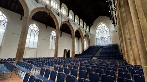 Shaun Whitmore/BBC Rows of blue chairs inside The Halls. In the background at the back of the chairs is a large stained glass window.