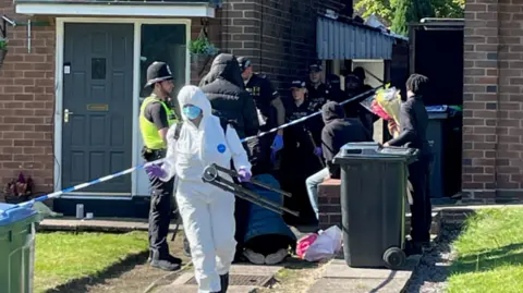 PA Images A group of police officers in black uniform and caps walk from the drive between two houses, with an officer in a white forensics suit, blue face mask and purple gloves stands at the front. The house is cordoned off by police tapes and an individual stands outside the cordon with a bunch of flowers, while a person in a blue coat kneels on the ground with their back to the camera.