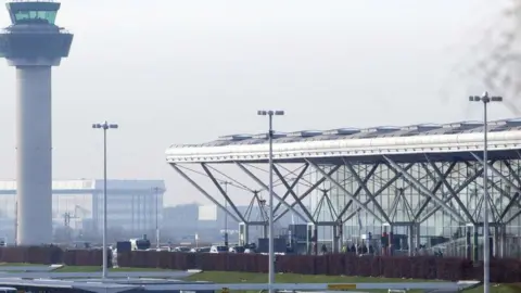 PA Media Stansted Airport, showing the tall control tower and white roof of the terminal building. Cars can be seen arriving at the drop-off at the terminal, with people entering the building.