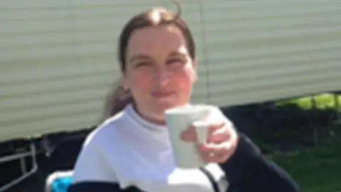 A woman with long brown hair in a ponytail smiles at the camera, holding up a white mug. She is outside and appears to be sitting in a camping chair, though it is only partially visible and the photograph is blurry.