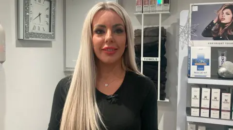 A woman in her 40s is pictured looking at the camera in a hairdressing salon. She is dressed in black and has long blonde hair.