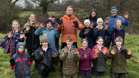 Thirteen children, their teachers, and naturalist Chris Packham show off their muddy hands. They are all dressed head-to-toe in waterproof clothing. 