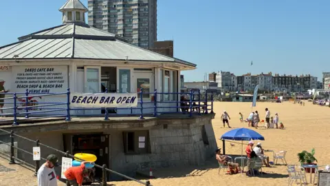 Simon Jones/BBC People relaxing at the Main Sands beach in Margate, Kent. A beach bar can be seen in the image, with Arlington House visible in the background. The skies are very blue.