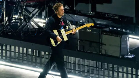 Sam Fender on stage with a black and white striped electric guitar. He's walking with a smile on his face.