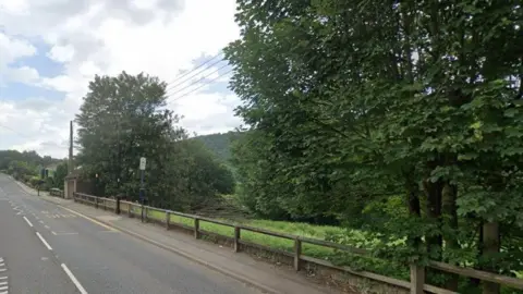 Google A picture looking up a road with housing to the left hand side and fields and trees to the right hand side.