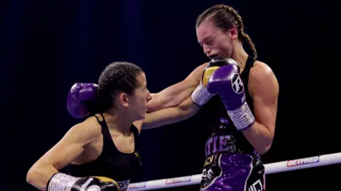 PA Media Nina Hughes hits opposing boxer Katie Healy during a fight. They are both wearing black vests and have their hair plaited. Healy is wincing as she takes the blow on her chin.