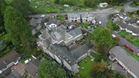 An aerial shot of the large home. It is grey-bricked, L-shaped with a lawn. There are large houses behind it and is near a road.