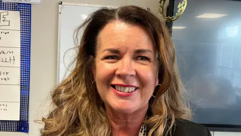 Jill West is smiling. She has dark wavy hair. She's wearing hoop earrings and has pink lipstick on.