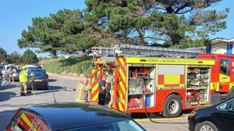 Dorset Police A fire engine is pictured parked across a road. Several firefighters can be seen working in the area. 