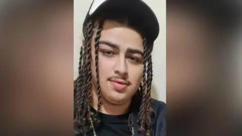 Northamptonshire Police Mason Miller had long dark brown hair in twisted plaits, and a moustache. He is wearing a black and white baseball hat, a black T-shirt and chain.