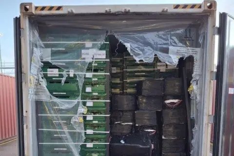 NCA A silver shipping container with the doors open. Inside are stacked green plastic trays and black packages wrapped in black plastic.
