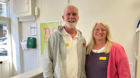 A couple in their 50s - a man with short grey hair and a beard and a woman with long blonde hair - smile as they stand in their office. The man is wearing a white polo shirt and a grey zip-up hoodie, while the woman is wearing a navy top and a pink cardigan.