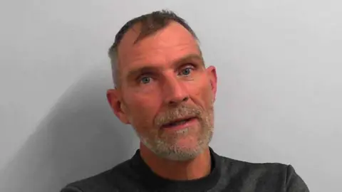 A custody picture of a man with short hair and a grey beard.